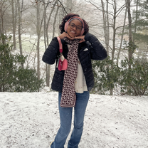 Smiling woman in glasses poses with hands under her chin in a snowy woodland, wearing a black coat, patterned scarf, jeans, and pink purse.