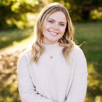 A young woman in a beige sweater smiles at the camera in a sunny outdoor setting, with greenery in the background.