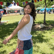 Smiling woman in a white sleeveless top and colorful patchwork skirt poses at a sunny outdoor festival, with bunting, tents, and vendors behind her.