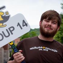Smiling bearded student holds a “Group 14” sign and decorated handle outdoors, wearing a Watagua College shirt; cheerful, upbeat mood.