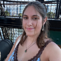 Smiling woman with brown hair and pearl earrings poses in a sunlit outdoor setting, wearing a lace-trimmed top and pendant necklace.