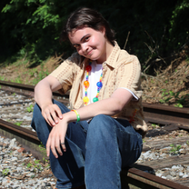 Smiling young person sits on railroad tracks outdoors, wearing a beige shirt, jeans, and colorful beaded necklaces.