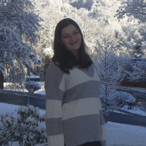 Young woman smiling gently in a gray-and-white striped sweater, standing outdoors amid snow-covered trees and a quiet winter landscape.