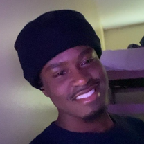 Close-up portrait of a smiling young man wearing a black knit beanie and shirt, lit with soft purple light indoors.