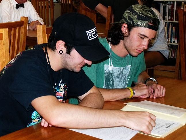 Two young men sit at a wooden table in a library, reviewing papers and reading together attentively.